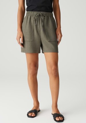 Frau trägt gerippte olivgrüne Shorts mit Kordelzug, schwarzen Top und schwarzen Slide-Sandalen, steht vor einem schlichten hellen Hintergrund.