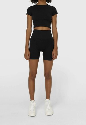 Woman wearing a black cropped short-sleeve top, high-waisted black biker shorts, and white lace-up ankle boots standing on a plain background.