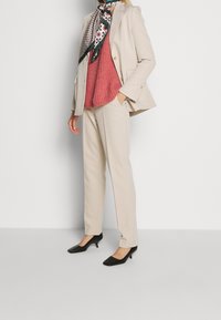 Giacca su misura beige con chiusura a bottoni dorati, abbinata a un top rosso texturizzato e tacchi neri a punta quadrata. La sciarpa presenta motivi geometrici.
