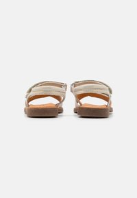 Vue arrière de sandales en cuir beige avec deux brides croisées, une semelle texturée et un talon plat. Les brides présentent une couleur claire et des détails de couture.