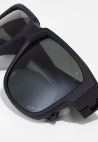 Ochelari de soare rectangulare negri cu lentile polaraizate și un finisaj mat. Prezintă un logo subtil pe lentilă și un design robust al ramei.