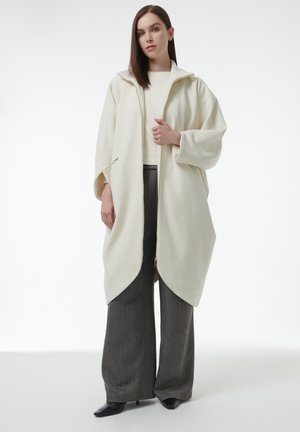 Crème-kleurige, gewatteerde jas met een rits aan de voorkant en een oversized ontwerp; gecombineerd met wijde, textuur grijze broek en zwarte puntige hakken.