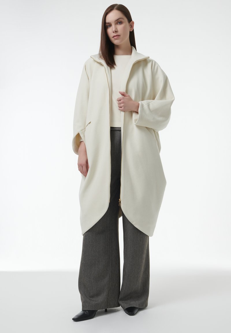 Abrigo de color crema con capucha, cierre de cremallera y diseño oversize; combinado con pantalones gris texturizados de pierna ancha y tacones negros de punta.