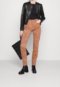 Chaqueta negra de cuero con hombreras acanaladas, combinada con pantalones naranjas con estampado y acabado texturizado, y botas negras de tacón.