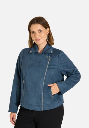 Frau mit kurzen braunen Haaren trägt eine blaue Jacke mit Reißverschluss und schwarze Hose, steht mit einer Hand in der Jackentasche vor weißem Hintergrund.