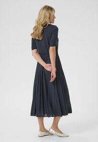 Marinblå, veckad klänning med korta ärmar; strukturerad tyg; cinchad midja. Silverfärgade ballerinas kompletterar outfitten. Bakvy visas.