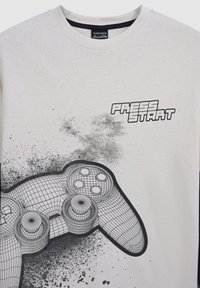 T-shirt in cotone color crema con una grafica di un controller di gioco in wireframe e il testo "PRESS START" in grassetto tipografia nera.