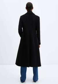 Cappotto di lana nero con un design lungo e fluente, colletto a vista e tasche laterali, abbinato a jeans a gamba larga blu.