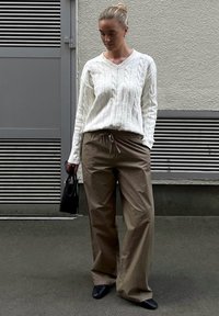 Weißer Kabelstrickpullover, lockere braune Hose mit elastischem Bund, schwarze Handtasche und schwarze Slippers. Glatte Texturen und neutrale Farben.