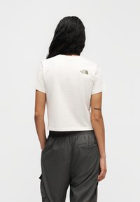 Κοντομάνικη cropped μπλούζα σε ανοιχτό μπεζ ύφασμα, με μικρό πράσινο λογότυπο στην πλάτη. Συνδυάζεται με παντελόνι σε σκούρο γκρι χρώμα με ελαστική μέση.