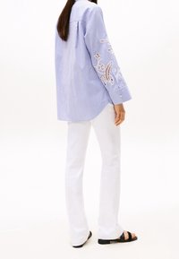 Camicia a maniche lunghe a righe azzurro chiaro e bianco con intricati ricami bianchi sulle maniche; abbinata a pantaloni bianchi a campana e sandali neri.