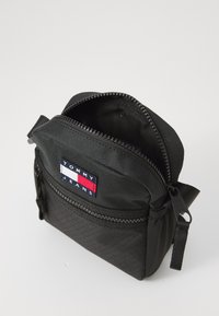 Sort nylon taske med struktureret overflade, et hovedrum med lynlås, en lynlåslomme foran og et Tommy Jeans mærke i rød, hvid og blå.