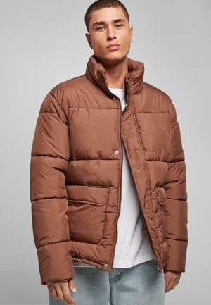 Brown padded jacket with a high collar, snap buttons, and a front pocket. Smooth texture and boxy shape, worn over a white shirt.