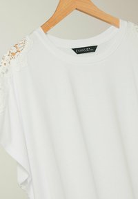 T-shirt bianca con scollatura rotonda, caratterizzata da dettagli in pizzo sulle spalle. Realizzata in tessuto morbido, appesa a un appendiabiti di legno.