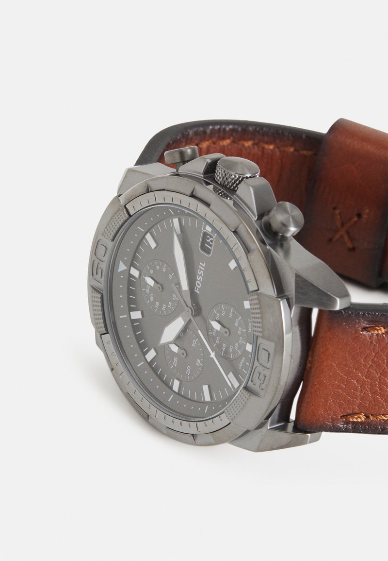 Montre chronographe grise avec un boîtier en métal, bracelet en cuir marron texturé et trois sous-cadrans sur un cadran foncé.