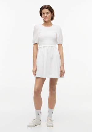 Młoda kobieta stoi ubrana w białą sukienkę z bufkami i marszczoną górą, białe skarpetki z logo oraz białe trampki na jednolitym tle.