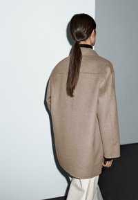 Oversized bruine wollen jas met een gladde textuur, klassieke kraag en verlaagde schouders, gecombineerd met lichtgekleurde broeken.