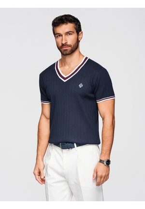 Mand iført marineblå kortærmet V-hals sweater med hvid og bordeaux kant, hvide bukser og et sølvur, stående mod en ensfarvet baggrund.