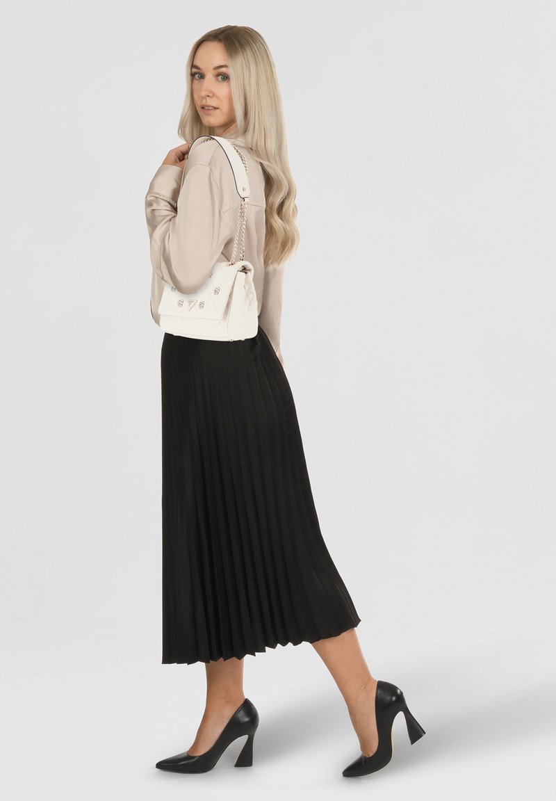 Bolso blanco acolchado con cadena y acentos metálicos, combinado con una blusa beige y una falda midi plisada negra, complementado con tacones negros.