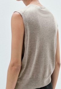 Knit top beige ilmanhihoilla, pyöreällä kauluksella ja pehmeällä tekstuurilla. Takana hieman rento malli ja hillitty röyhelö.