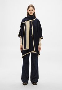 Πόντσο σε ναυτικό μπλε και εκρού με κούμπωμα με κουμπιά, αντίθετη επένδυση και μαλακό ύφασμα. Συνδυάζεται με παντελόνι τζιν σε σκούρο χρώμα με φαρδιά μπατζάκια.