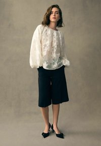 Chemisier blanc texturé avec des motifs floraux, associé à un pantalon noir court. Le modèle porte des escarpins noirs avec des nœuds, se tenant devant un fond neutre.