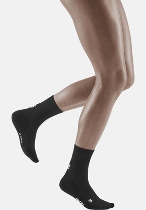 Schwarze Sportsocken aus strukturiertem Stoff, mit einem gerippten Bund und weißem Branding an der Seite, abgebildet an Beinen in Bewegung.