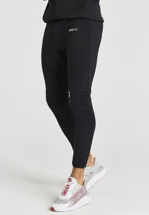 Jogging en noir avec une coupe slim, présentant une texture lisse et un petit logo blanc sur la cuisse. Assortis avec des baskets grises et rouges.