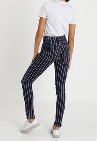 Jeans de mezclilla oscuros de tiro alto con rayas verticales rojas, que presentan un diseño ajustado y bolsillos traseros estándar. Combinados con zapatillas blancas.