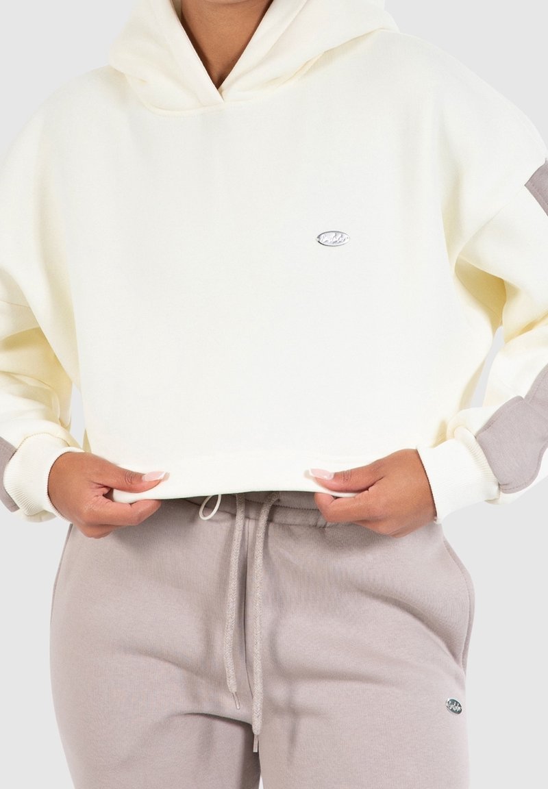 Personne portant un hoodie court crème avec des patches gris et un pantalon de survêtement gris à cordon, tenant l'ourlet du hoodie.