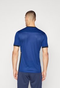 Sininen urheilupaita lyhyillä hihoilla, mustilla yksityiskohdilla kauluksessa ja sivuilla, sileä tekstuuri ja istuva muotoilu, nähtynä takaa.