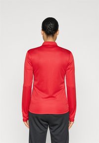 Κόκκινο μακρυμάνικο τοπ από λείο, ελαστικό ύφασμα με γιακά. Η πλάτη έχει εφαρμοστό σχέδιο και διακριτικές ραφές. Μαύρο παντελόνι φορεμένο από κάτω.