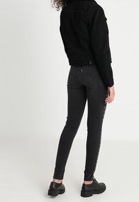 Czarna kurtka dżinsowa o klasycznym kroju, w połączeniu z dopasowanymi czarnymi dżinsami z bocznymi guzikami. Tekstura wydaje się gładka i elastyczna.