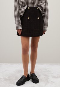 Minifalda de tweed negro con botones dorados, combinada con una camisa de cuadros de manga larga y mocasines negros de suela gruesa. Tejido suave y texturizado.