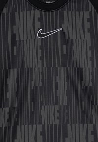 Camisa atlética negra con tejido texturizado y logotipos de Nike verticales de diferentes tamaños. Acentuada por un logotipo de swoosh de Nike en blanco.