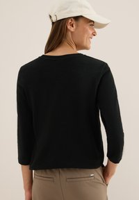 Femme de dos portant un haut à manches longues noir, un pantalon beige et une casquette blanche, souriant devant un fond clair.