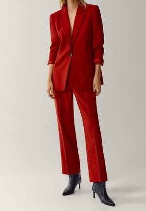 Femme portant un tailleur rouge ajusté avec un blazer à un seul bouton et un pantalon large assorti, associé à des bottines noires à talons.