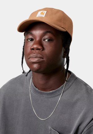 Persona joven de piel oscura y cabello trenzado que lleva una gorra beige, camiseta gris de cuello redondo y un collar delgado de cadena plateada, con expresión neutra.