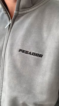 Casaco cinza com fecho éclair, feito de tecido ribado, com um colarinho alto e o logótipo bordado em preto "PEGADOR" no lado esquerdo.