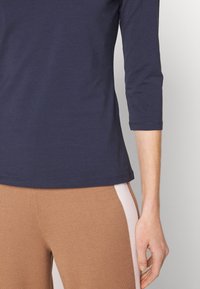 Top blu Navy con maniche a tre quarti realizzato in tessuto morbido, vestibilità leggermente rilassata e texture liscia. Abbinato a pantaloni marroni a coste con dettagli chiari.