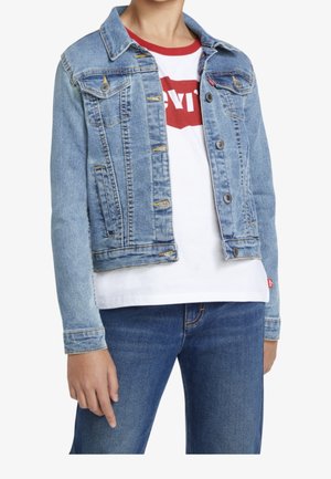 Persoon die een lichtblauwe spijkerjas draagt over een wit T-shirt met rood logo en een blauwe spijkerbroek, staand met de linkerhand ontspannen langs het lichaam.