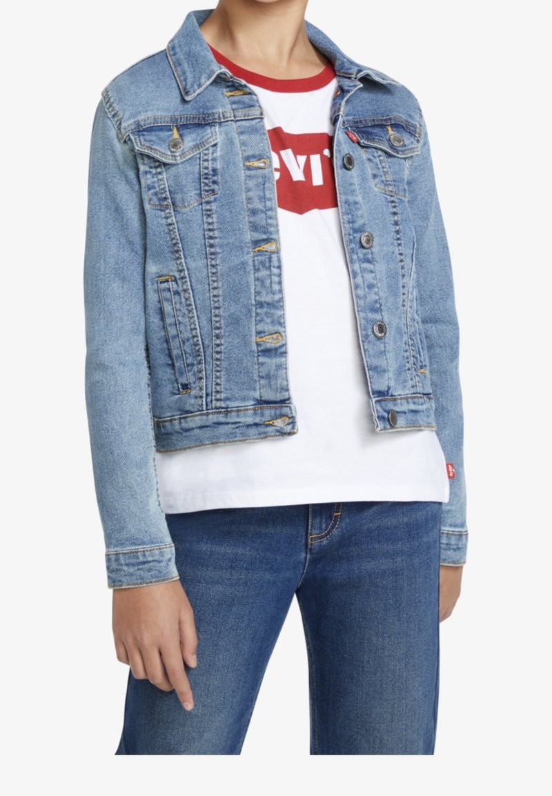 Personne portant une veste en jean bleu clair par-dessus un T-shirt blanc avec un logo rouge et un jean bleu, debout avec la main gauche détendue sur le côté.
