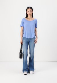 Cămașă scurtă de culoare albastru deschis, cu guler larg, purtată cu blugi evazați din denim și adidași albi. Ținând o geantă mică de mână, neagră.