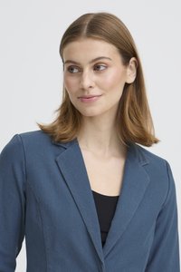 Noor naine, kellel on sirged helepruunid juuksed, seljas sinine jakk musta topi kohal, vaatab vasakule ühevärvilise tausta ees.