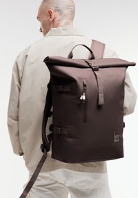 Mochila marrón de tipo roll-top con correas ajustables, hecha de un material texturizado. Cuenta con un bolsillo frontal con cremallera y una marca en el lateral.