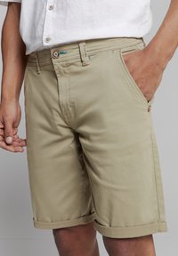 Beige katoenen shorts met een opgerolde zoom, zijzakken en een knoopsluiting met turquoise stiksels. Soepele textuur en casual ontwerp.