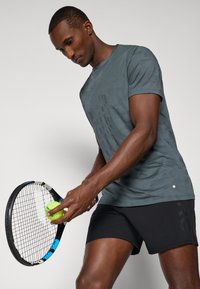 Γκρι αθλητικό μπλουζάκι με διακριτό σχέδιο, συνδυασμένο με μαύρες σορτς. Το άτομο κρατάει μία ρακέτα τένις και μία νεον πράσινη μπάλα.