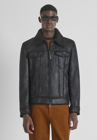 Man in een zwarte schapenvachtjas met rits en klepzakken, bruine corduroy broek en zwarte rechthoekige zonnebril.