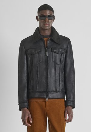 Man wearing black shearling jacket with zipper and flap pockets, brown corduroy pants, and black rectangular sunglasses.