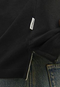Mustapaita, jossa on näkyvä valkoinen "ORIGINALS" -label. Kangas näyttää sileältä ja se on osittain päällekkäin haalistuneen denimkankaan kanssa.